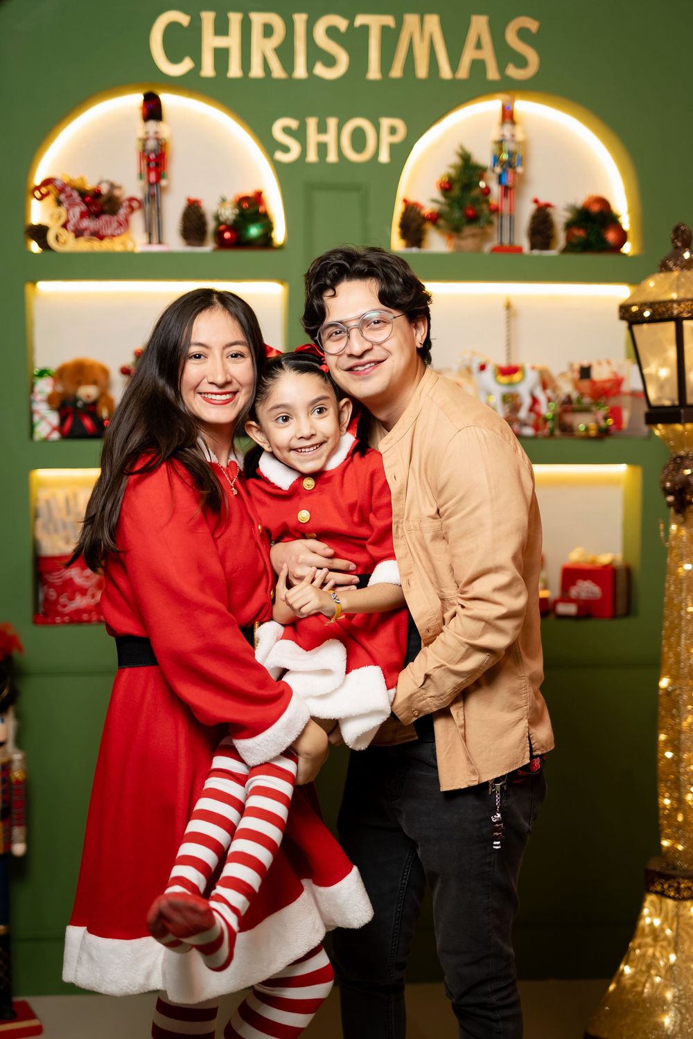 A festive Christmas photo session with family members wearing red and tan outfits against a green backdrop with lights.
