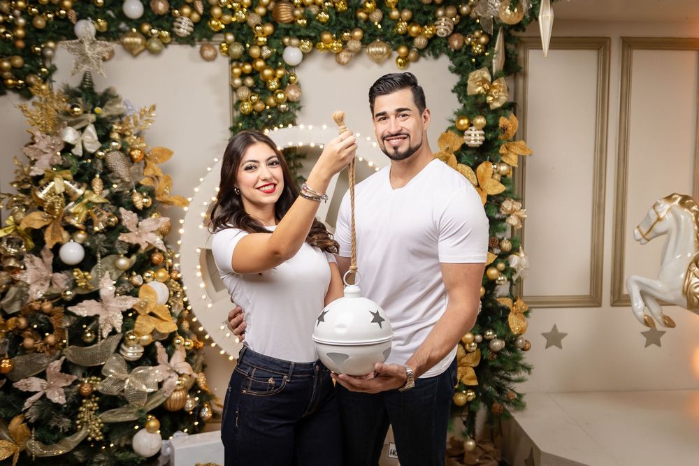 A couple in white and black outfits poses in front of a decorated gold Christmas tree and festive holiday decor.