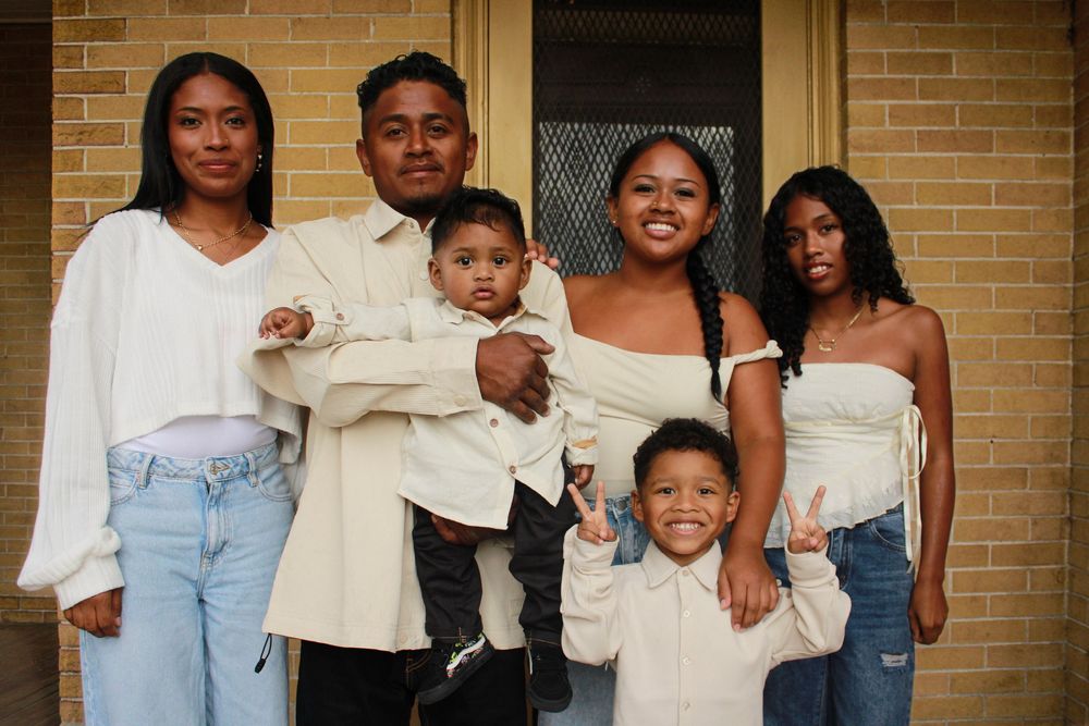 A happy family wearing coordinated outfits in light blue jeans and white tops poses together against a brick wall.