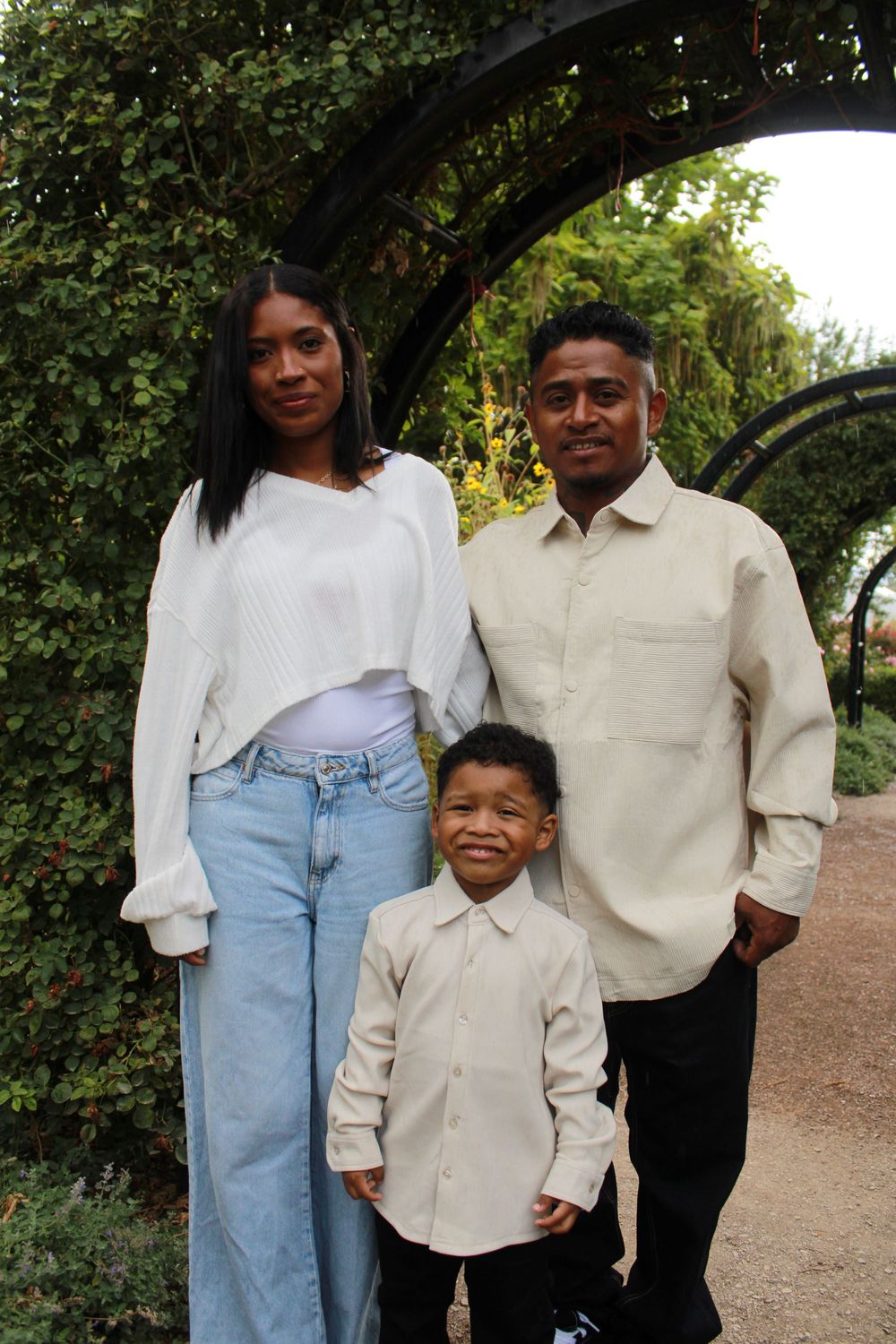 A family of three wearing light colored clothing poses together on a path surrounded by greenery.