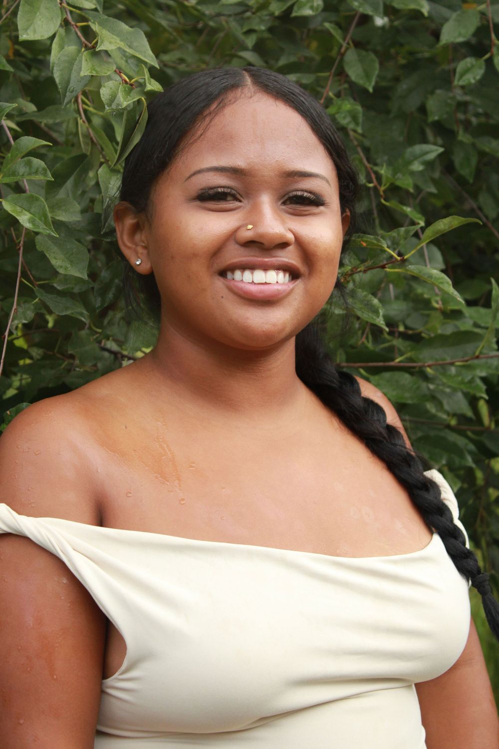 Person in cream-colored off-shoulder top smiles warmly against green foliage background.