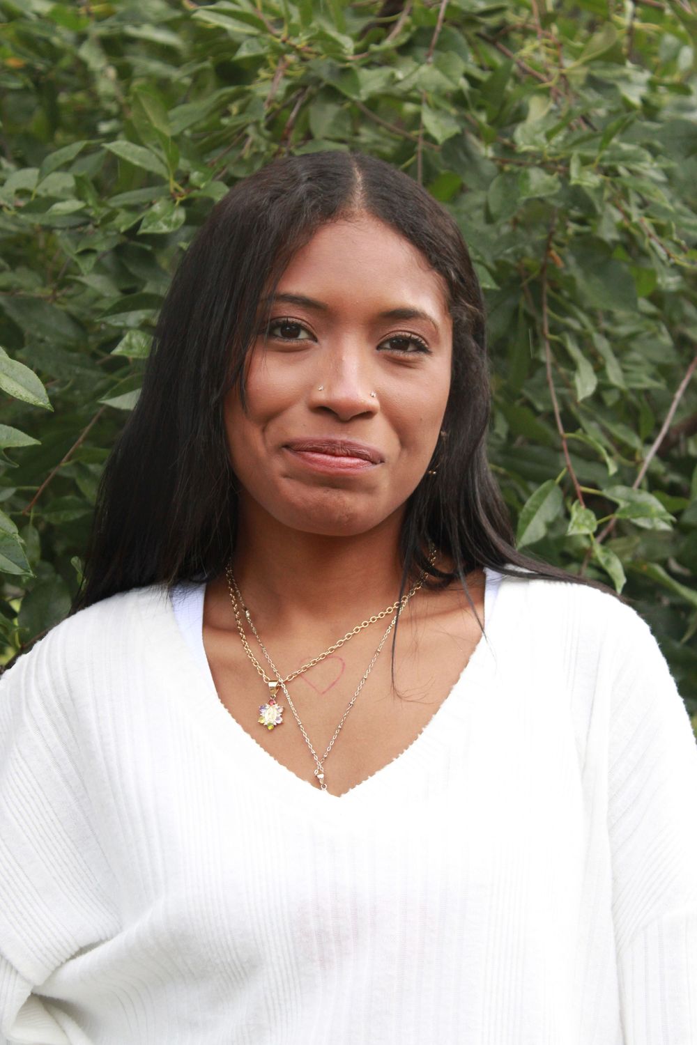 A person in a white sweater poses outdoors against a leafy green background.