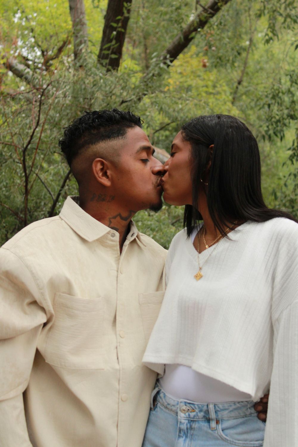 A couple shares a romantic kiss in a lush green forest setting while wearing coordinating cream-colored outfits.