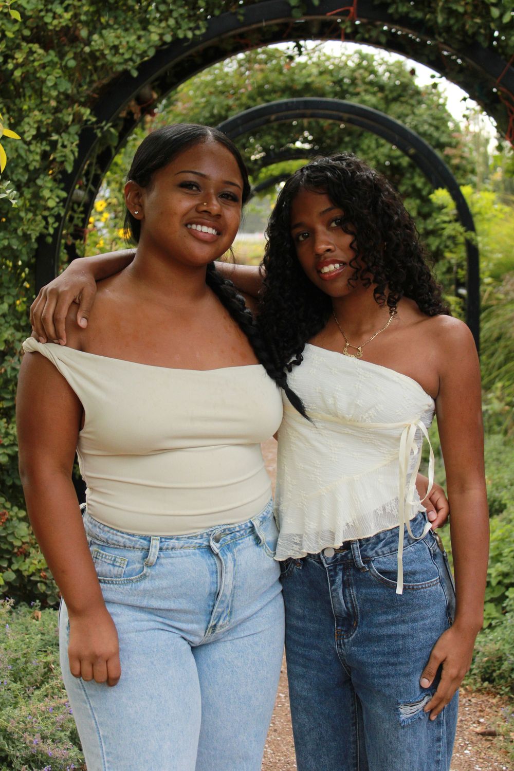 Two friends wearing white tops and blue jeans share joyful laughter while posing together outdoors in a garden setting.