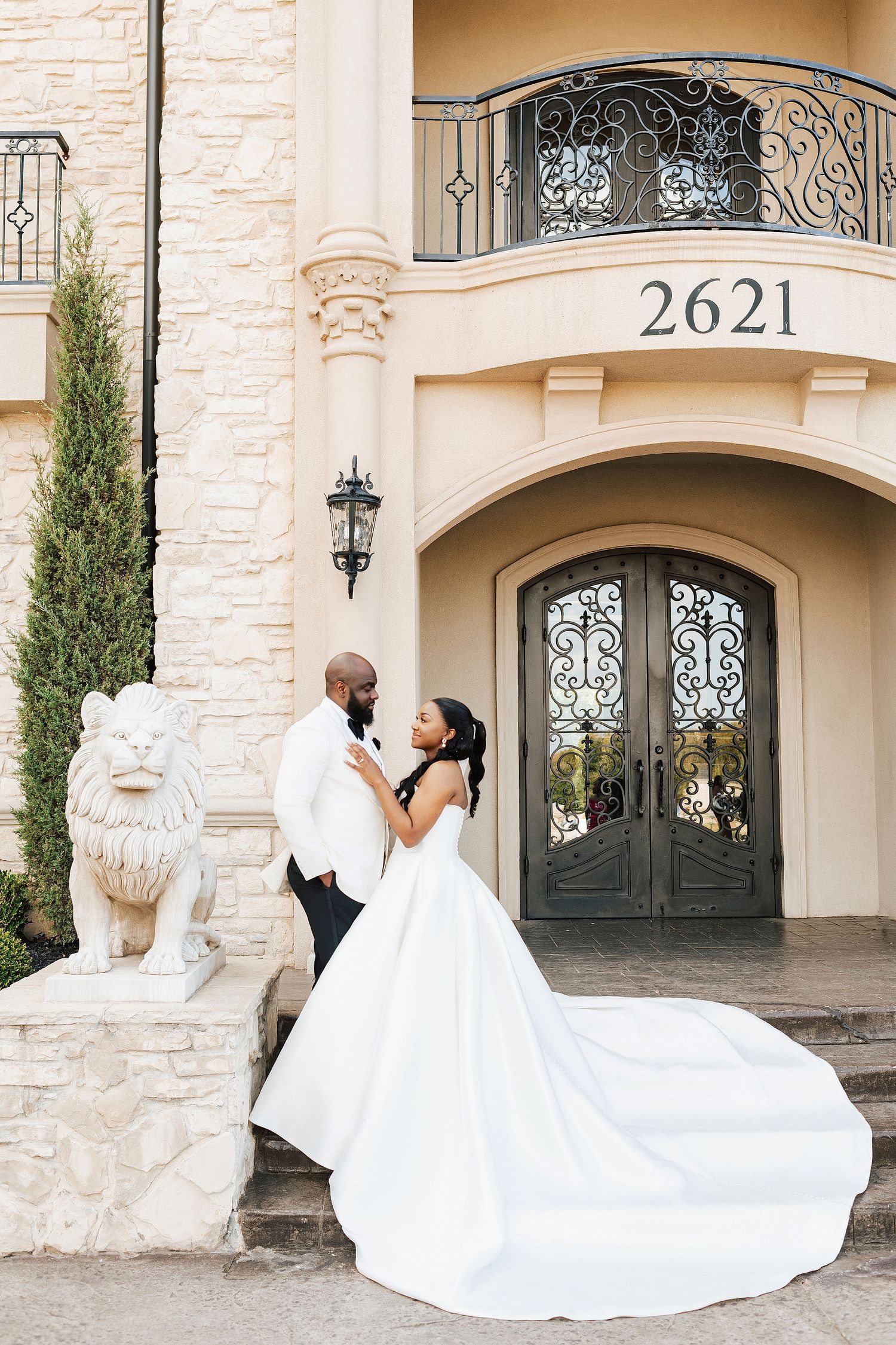 Elegant wedding couple poses in front of a luxurious stone mansion with ornate iron doors and decorative lion statues.