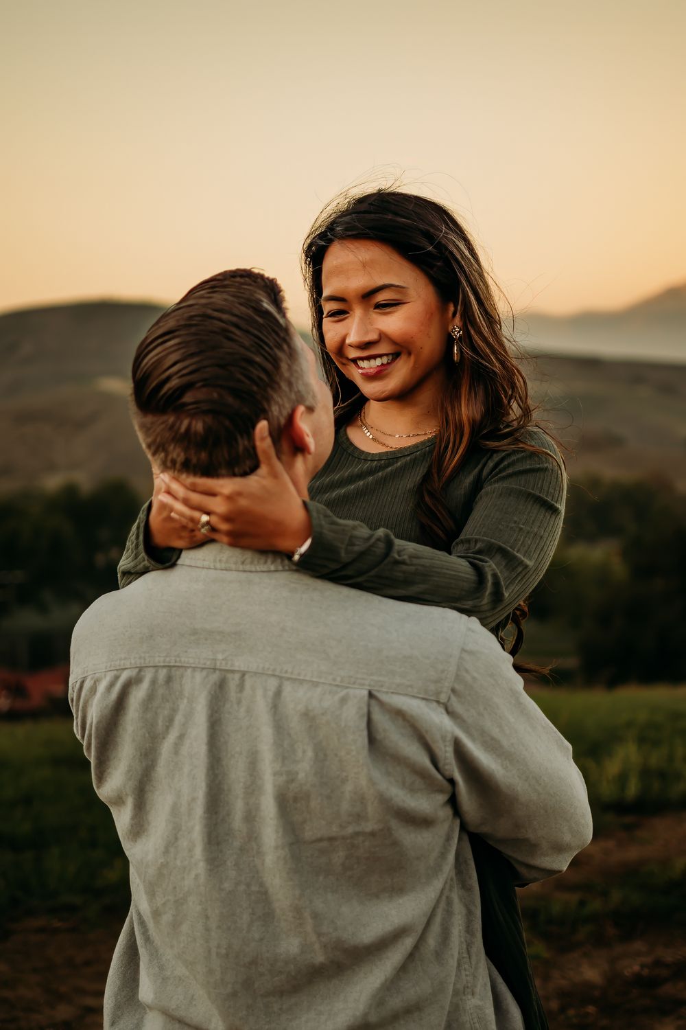 A smiling woman embraces a man outdoors at sunset with rolling hills in the background.