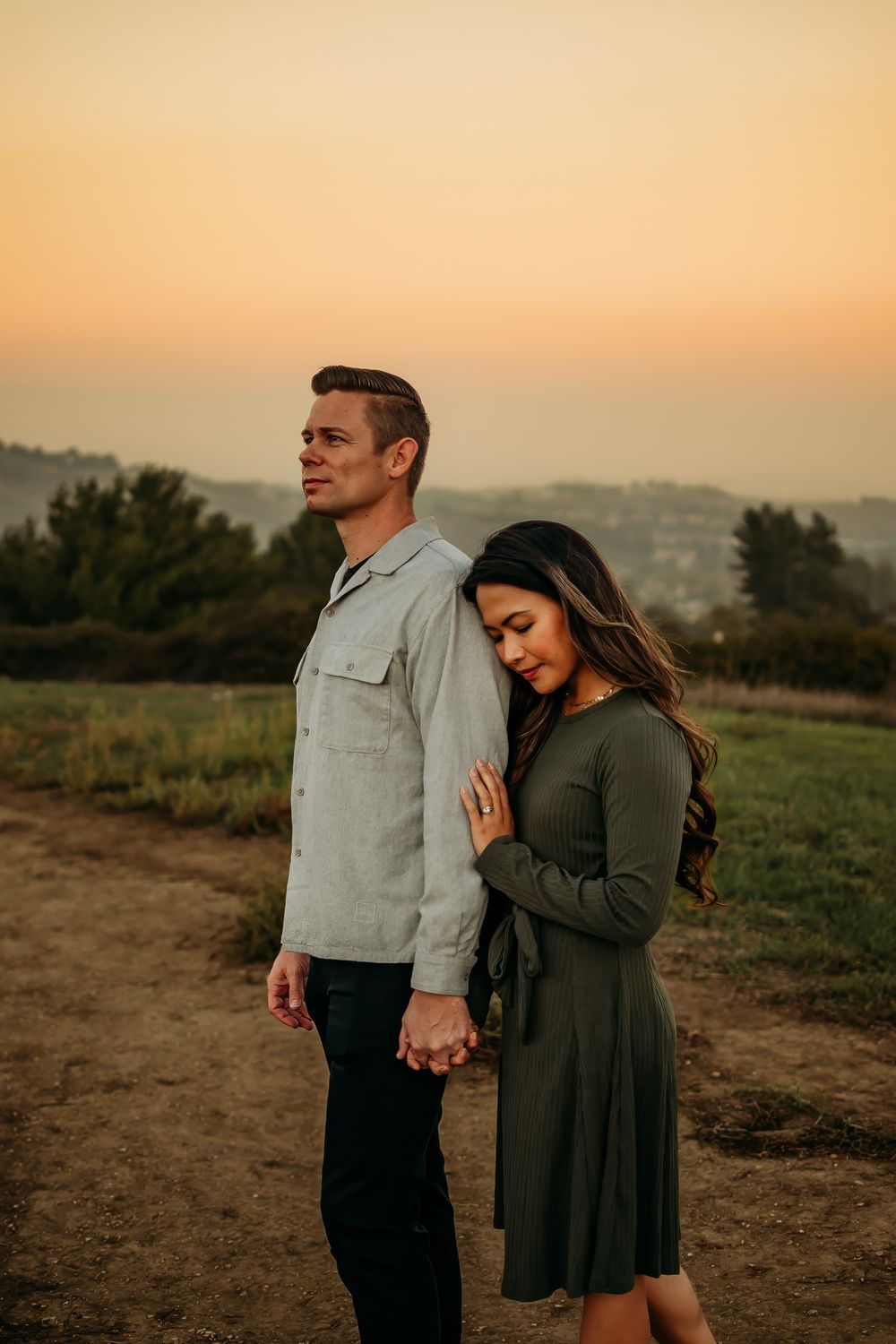 Couple holding hands at sunset in a field, woman in green dress leaning on mans back, golden hour sky.