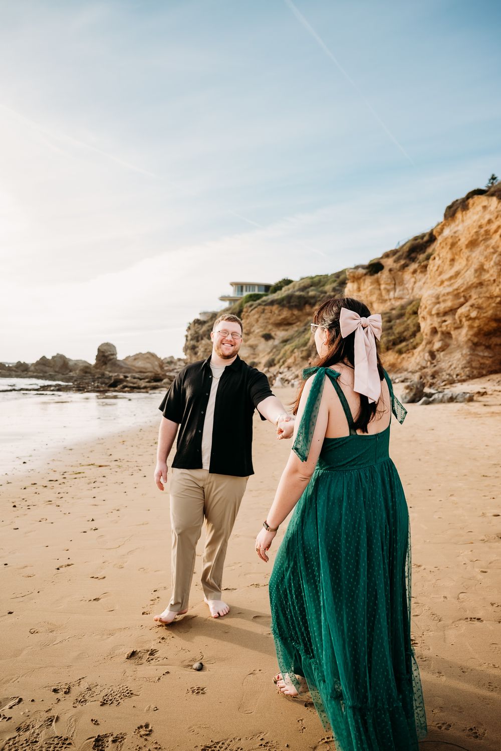 Couple walks playfully along sandy beach with cliffs in background.