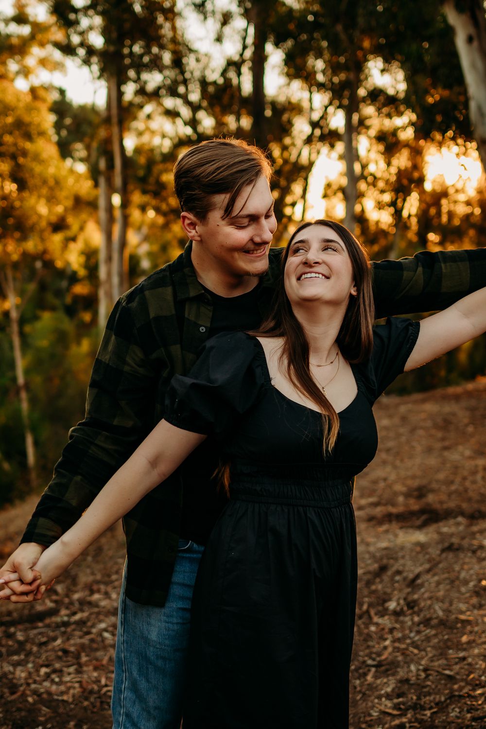 Playful couple laughs and dances together in a wooded area during golden hour.