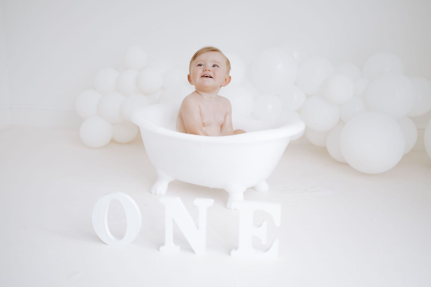 Baby in white onesie sits on white backdrop surrounded by soft white balloon decorations for studio photography.