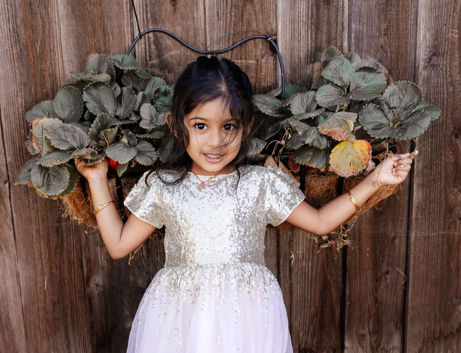 A girl wearing a white dress smiles in front of her strawberry plant in their Bay Area garden