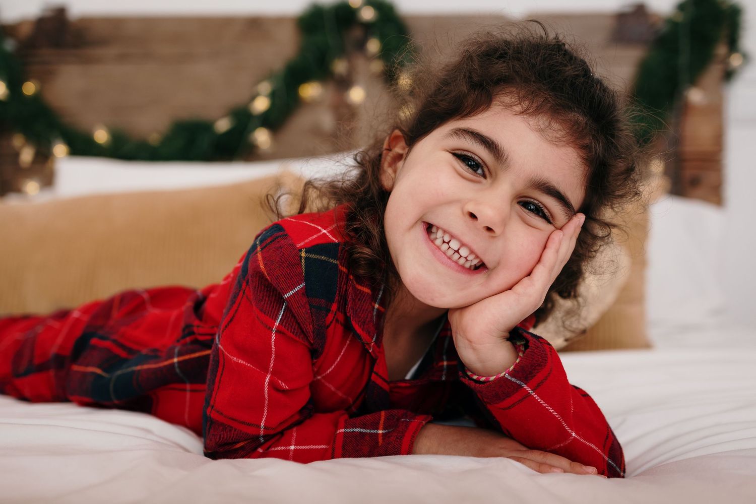 Child in red plaid pajamas poses playfully on white bedding.
