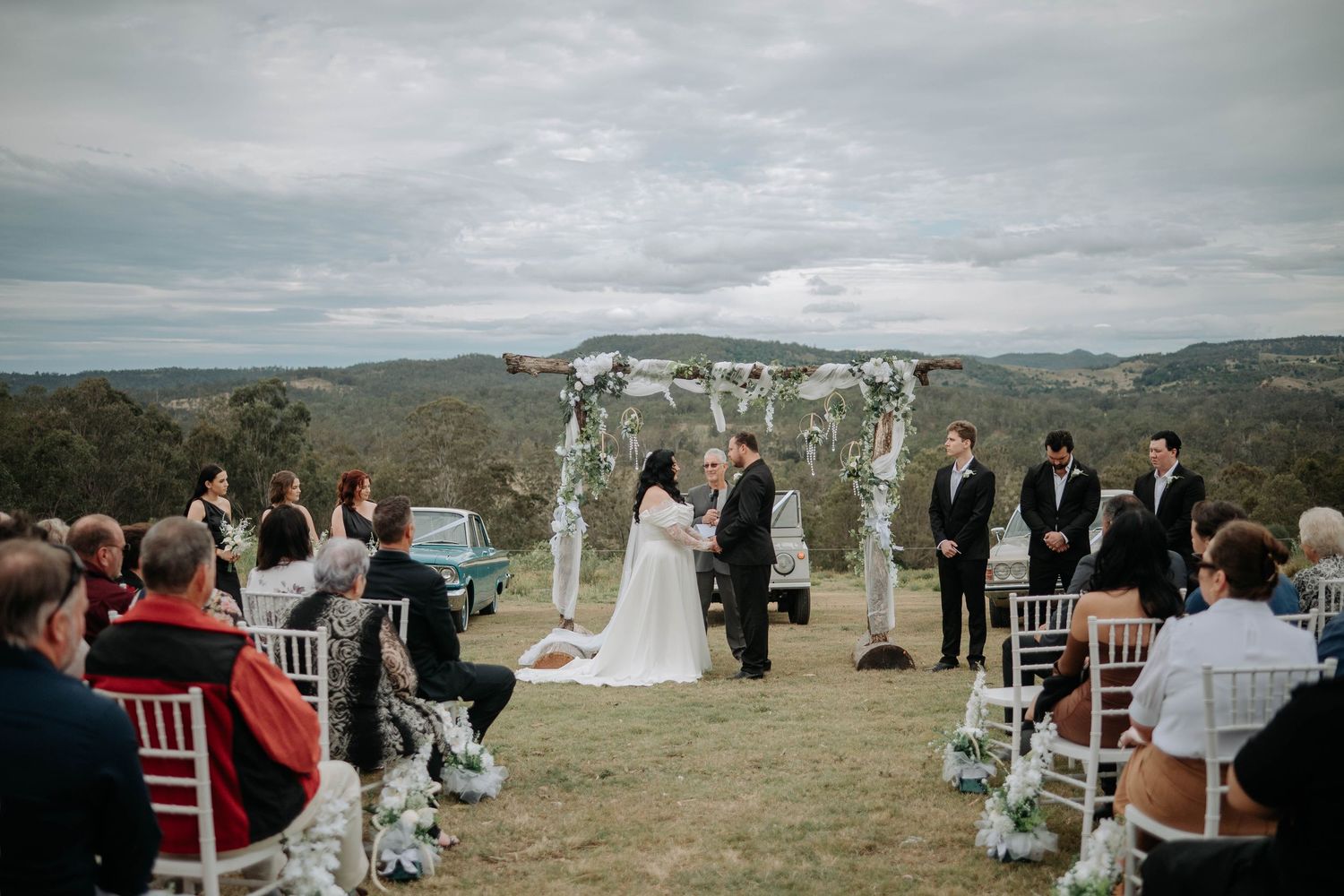 A panoramic view of an outdoor wedding ceremony set against rolling hills with guests seated in white chairs.