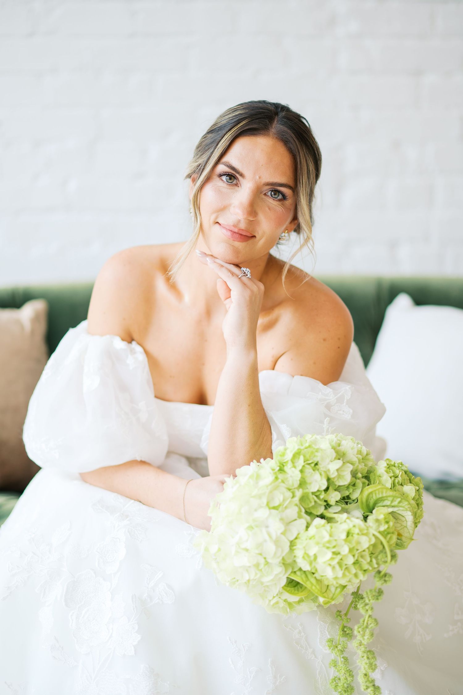 Elegant bridal portrait with soft lighting and green hydrangea bouquet on a plush sofa.