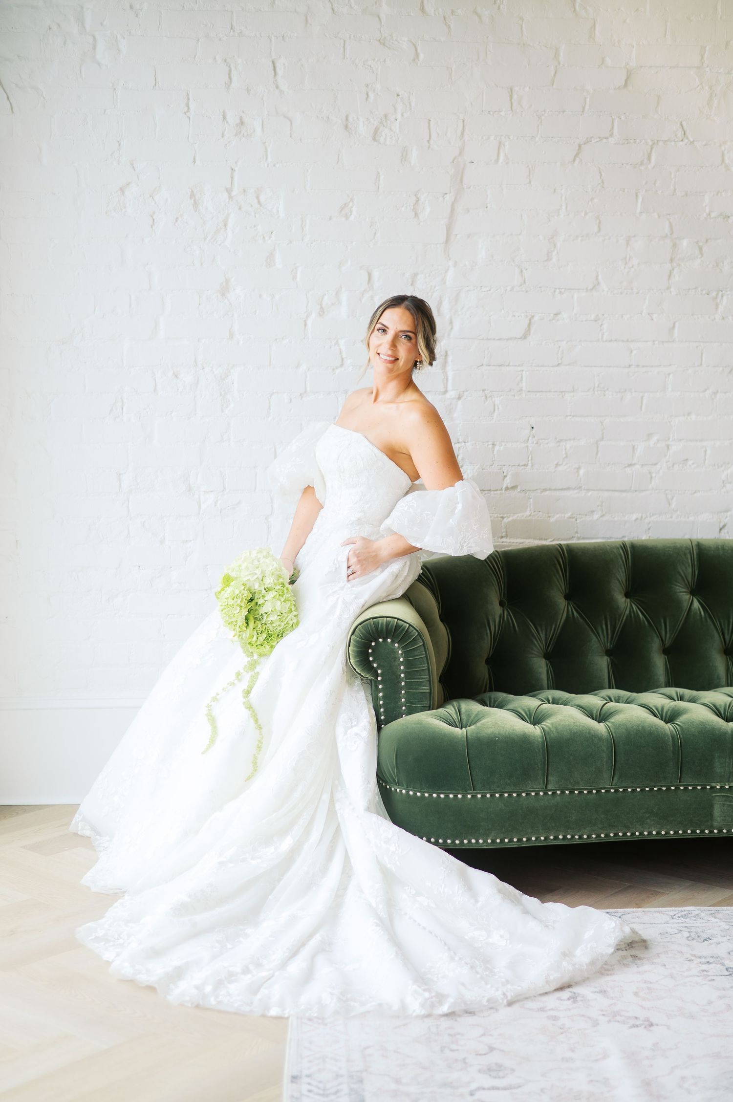 A bride in a white lace wedding gown poses elegantly on a dark green velvet tufted sofa against a white brick wall.