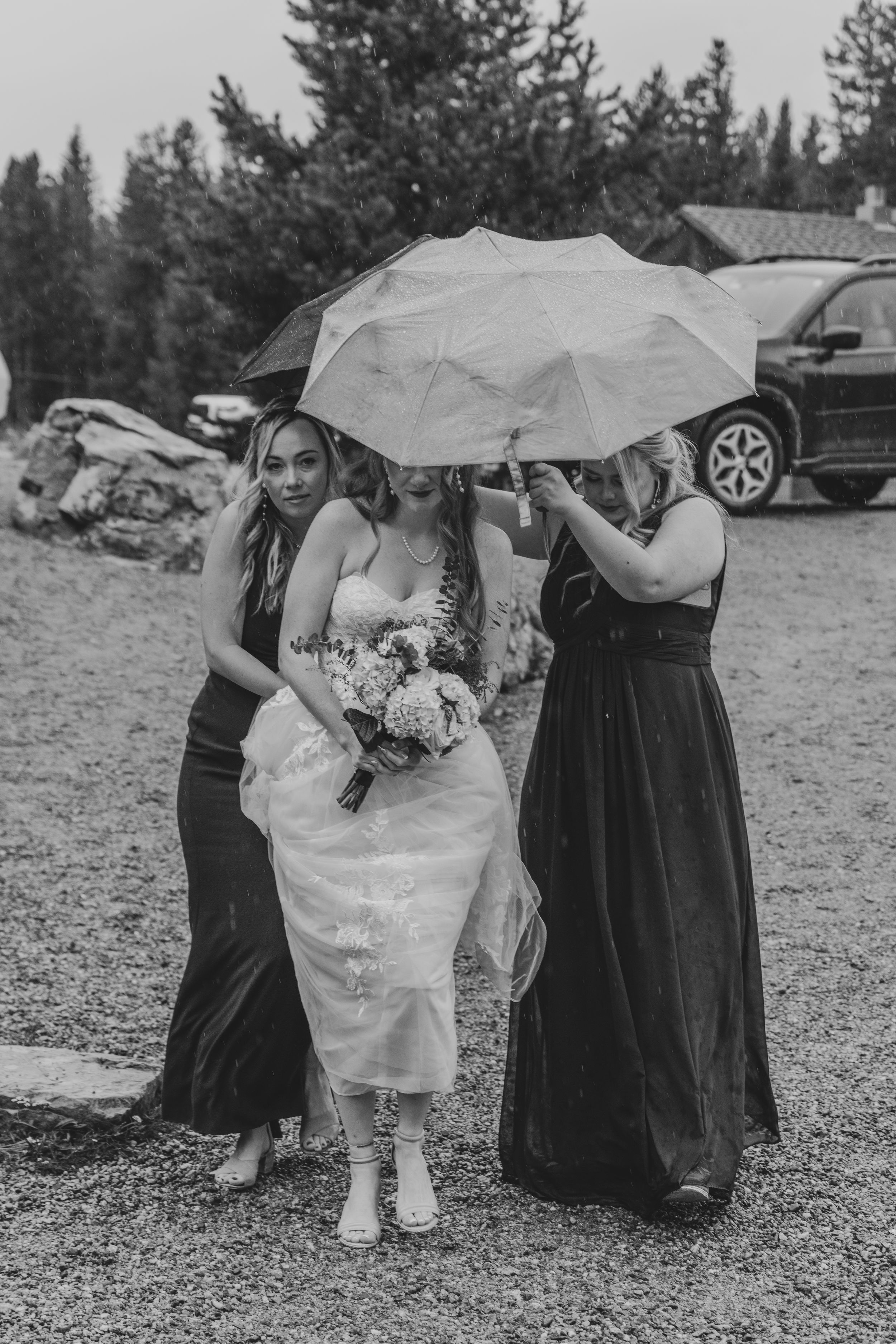 A group in formal attire huddles under white umbrellas on a gravel surface during an outdoor event.
