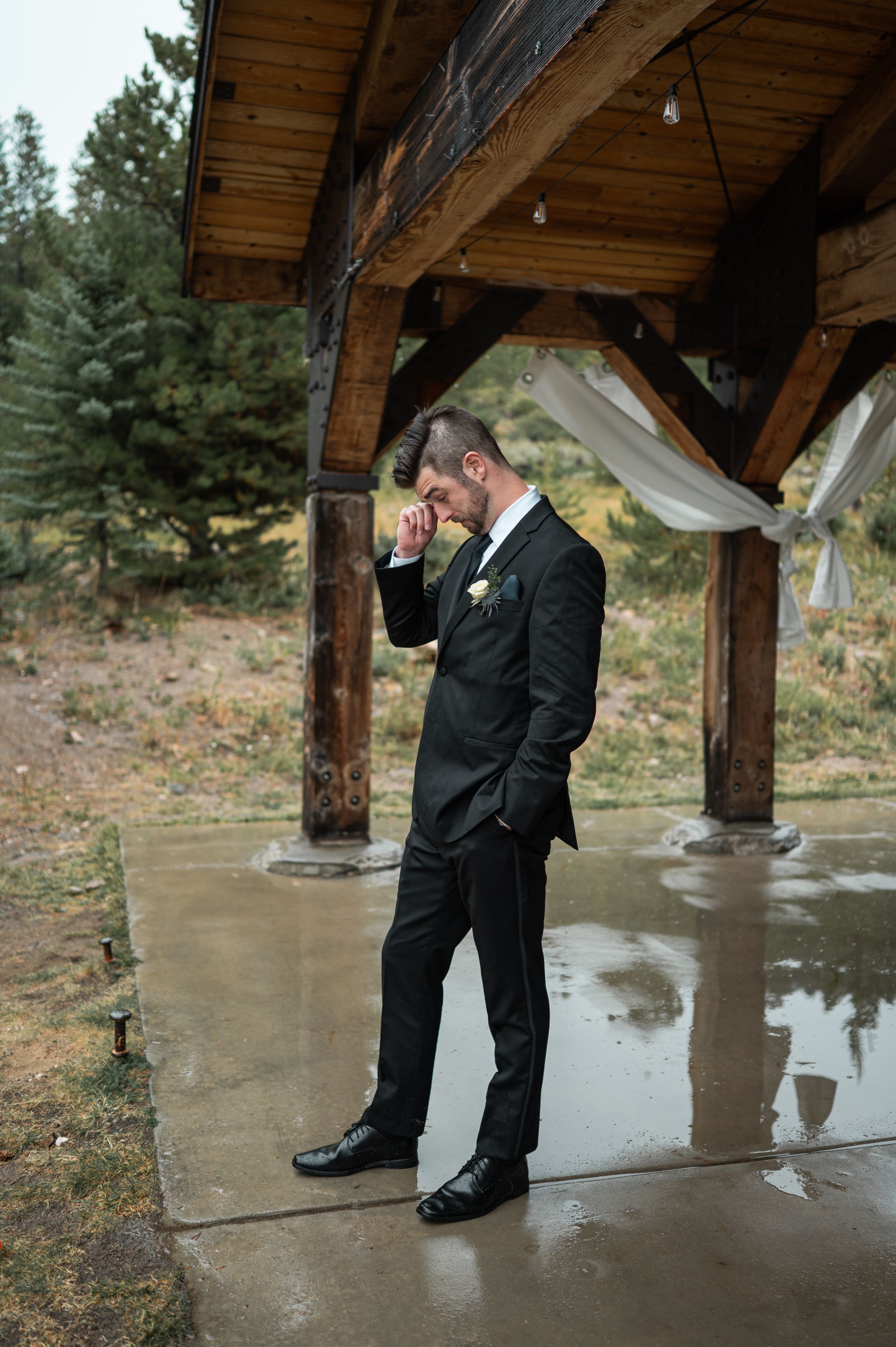 A groom in a black tuxedo stands under a rustic wooden pavilion during a rainy wedding day.