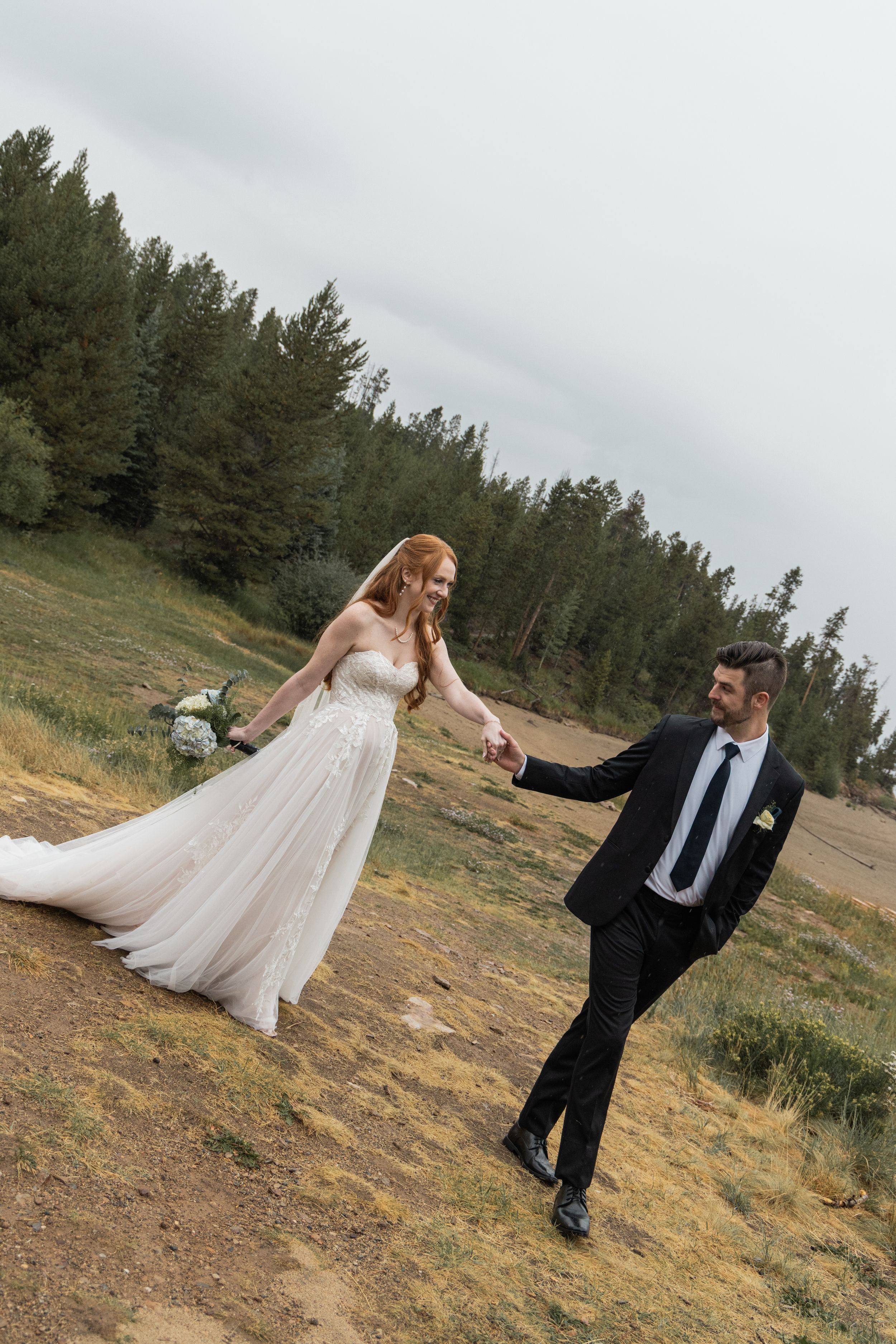 Playful wedding couple poses dramatically on a hillside with evergreen trees in the background.
