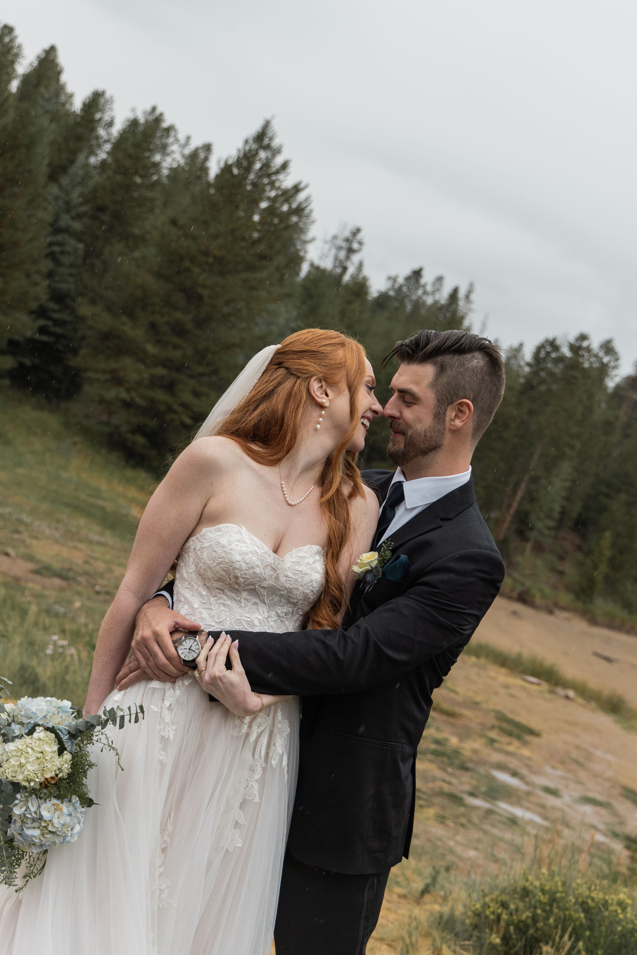 Romantic newlyweds share an intimate moment in a rustic outdoor setting with pine trees.