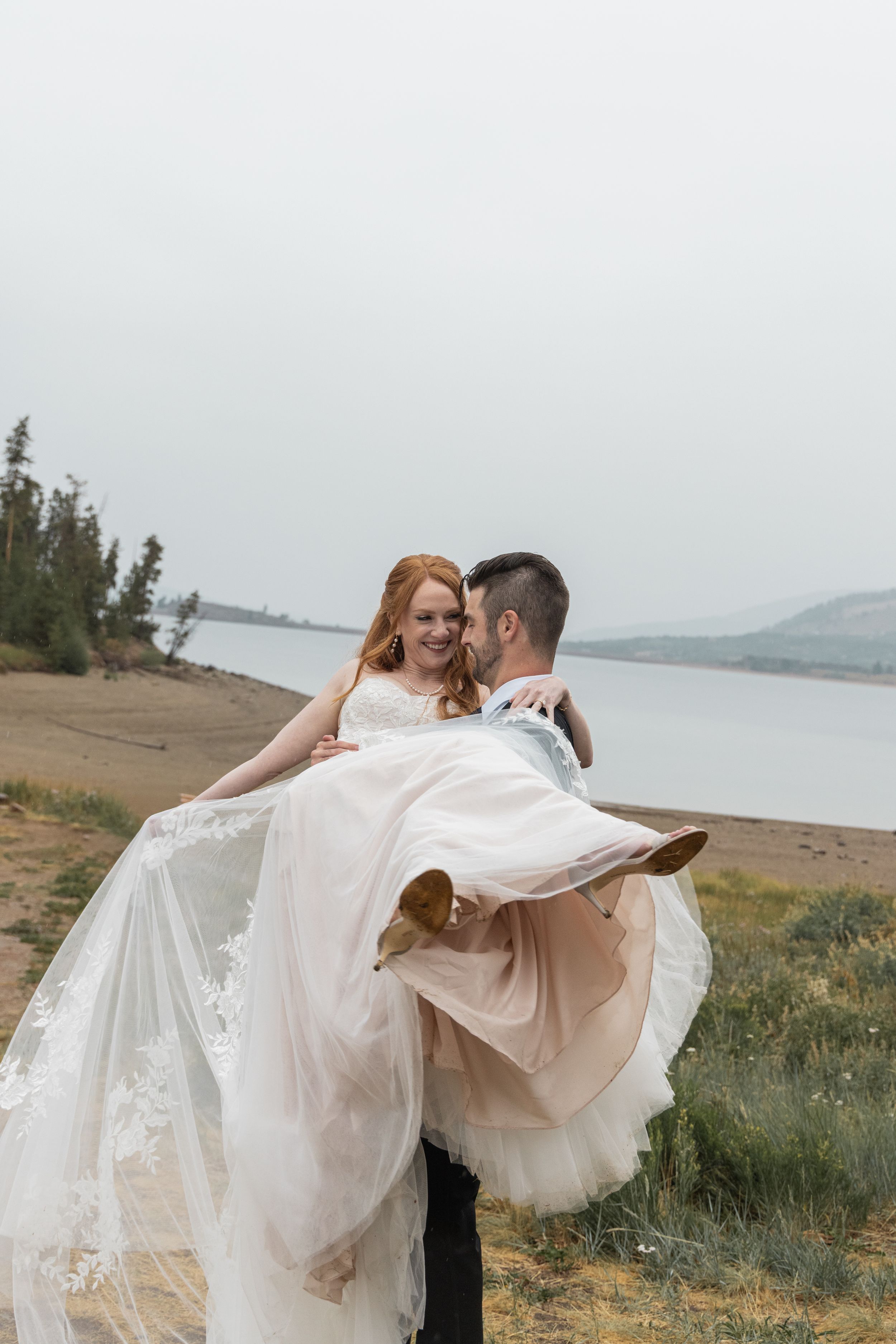 Flowing white wedding dress billows dramatically against a scenic mountain lake backdrop.