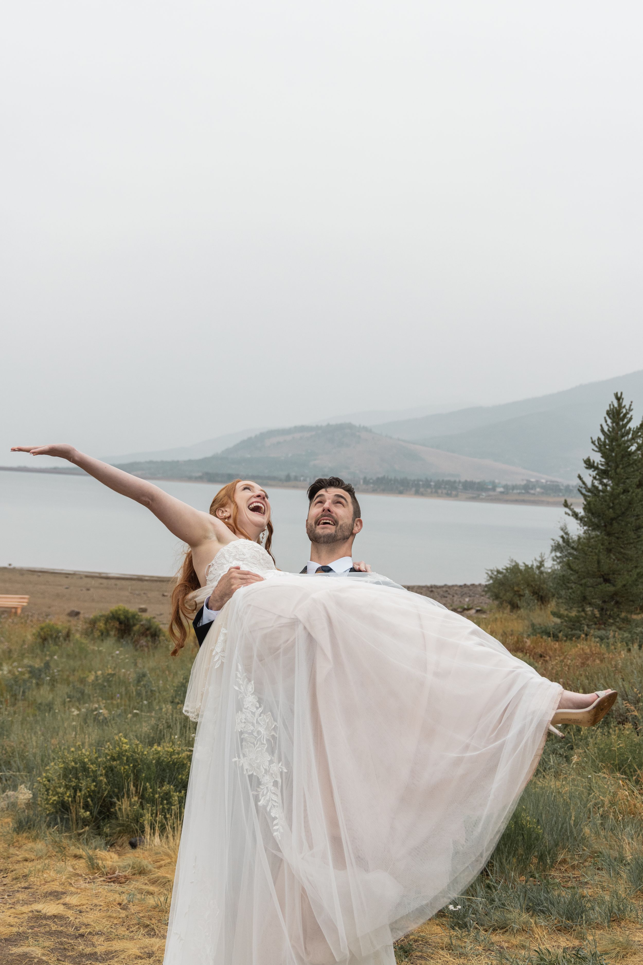 A couple in wedding attire dances joyfully in a scenic mountain meadow with water views in the background.