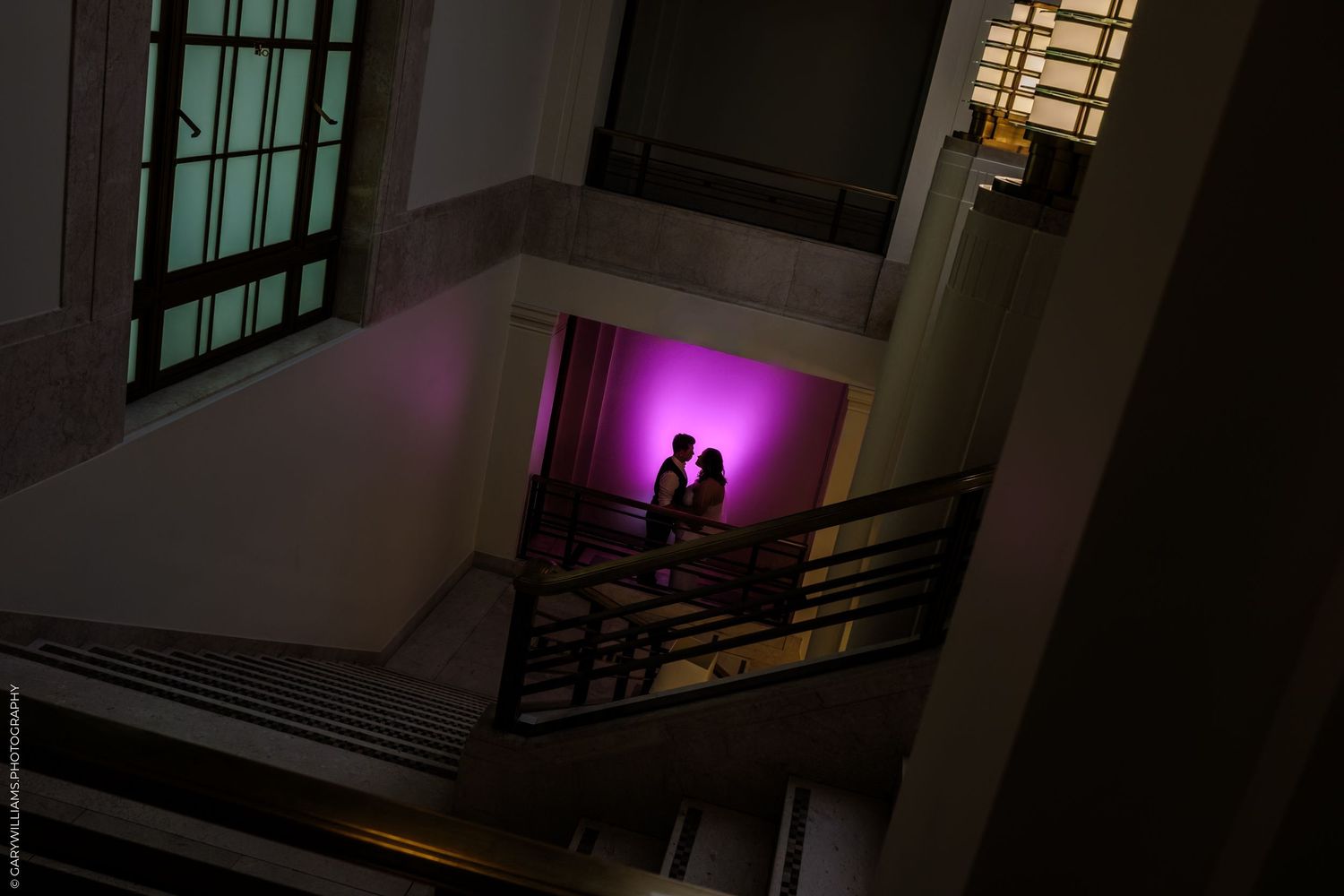Silhouettes stand on a staircase landing against a vibrant purple-lit wall in a dark interior space.