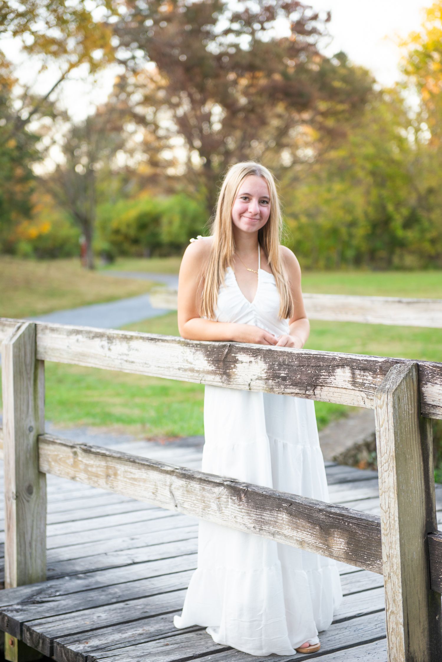 A person in a white dress poses on a rustic wooden boardwalk surrounded by autumn trees at sunset.