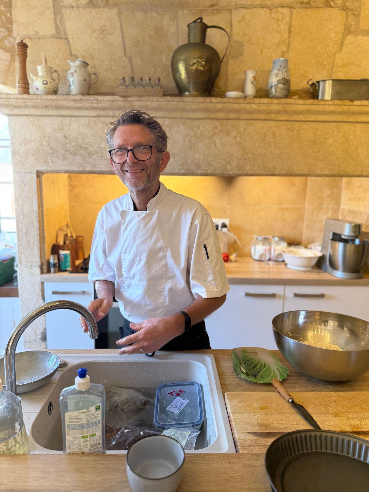 Chef in white uniform working at kitchen sink with vintage decor and pottery displayed on stone shelf above.