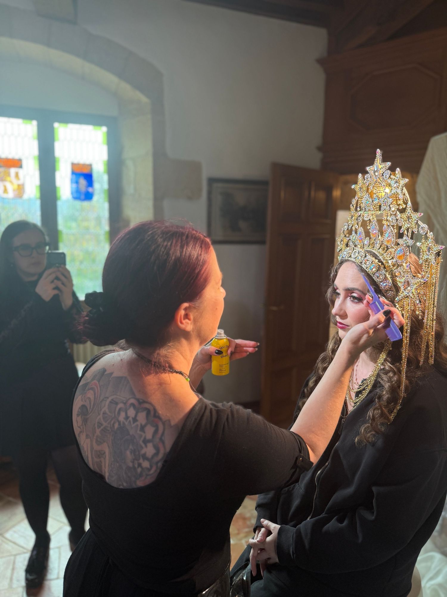 Makeup artist applying cosmetics on model wearing ornate crown in historic interior setting.