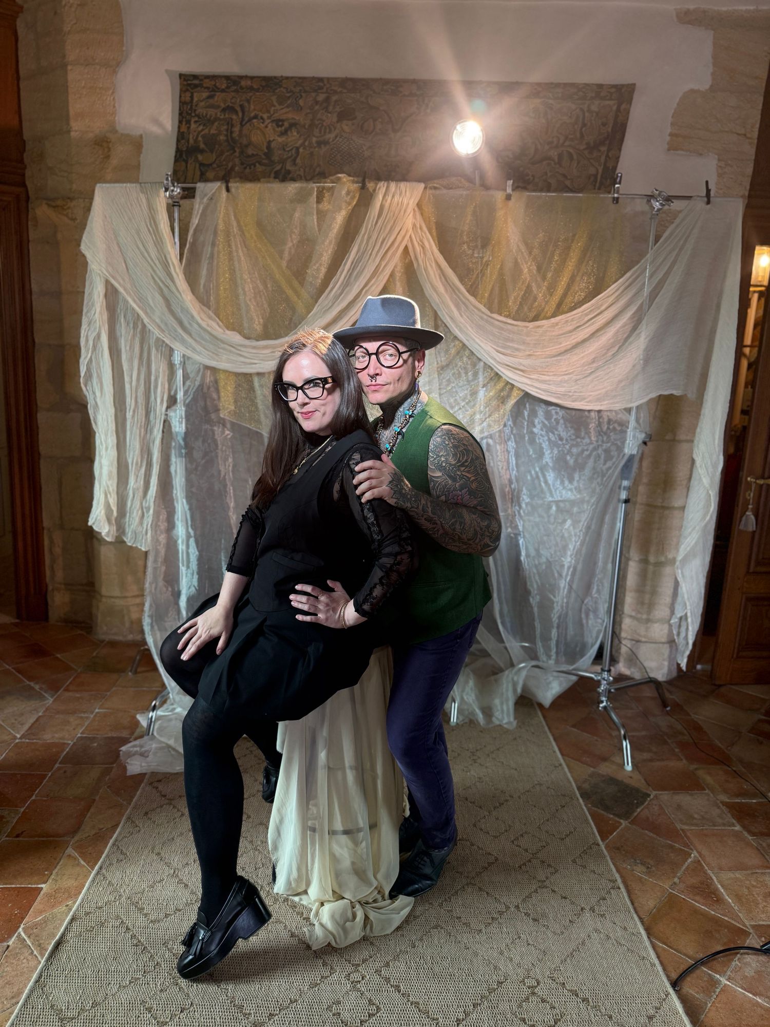 Couple posing playfully against draped fabric backdrop in vintage-style setting.