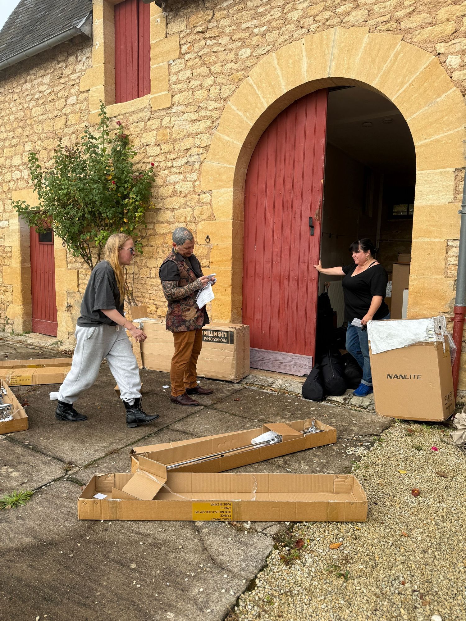 People assembling furniture from cardboard boxes outside a stone building with red doors.