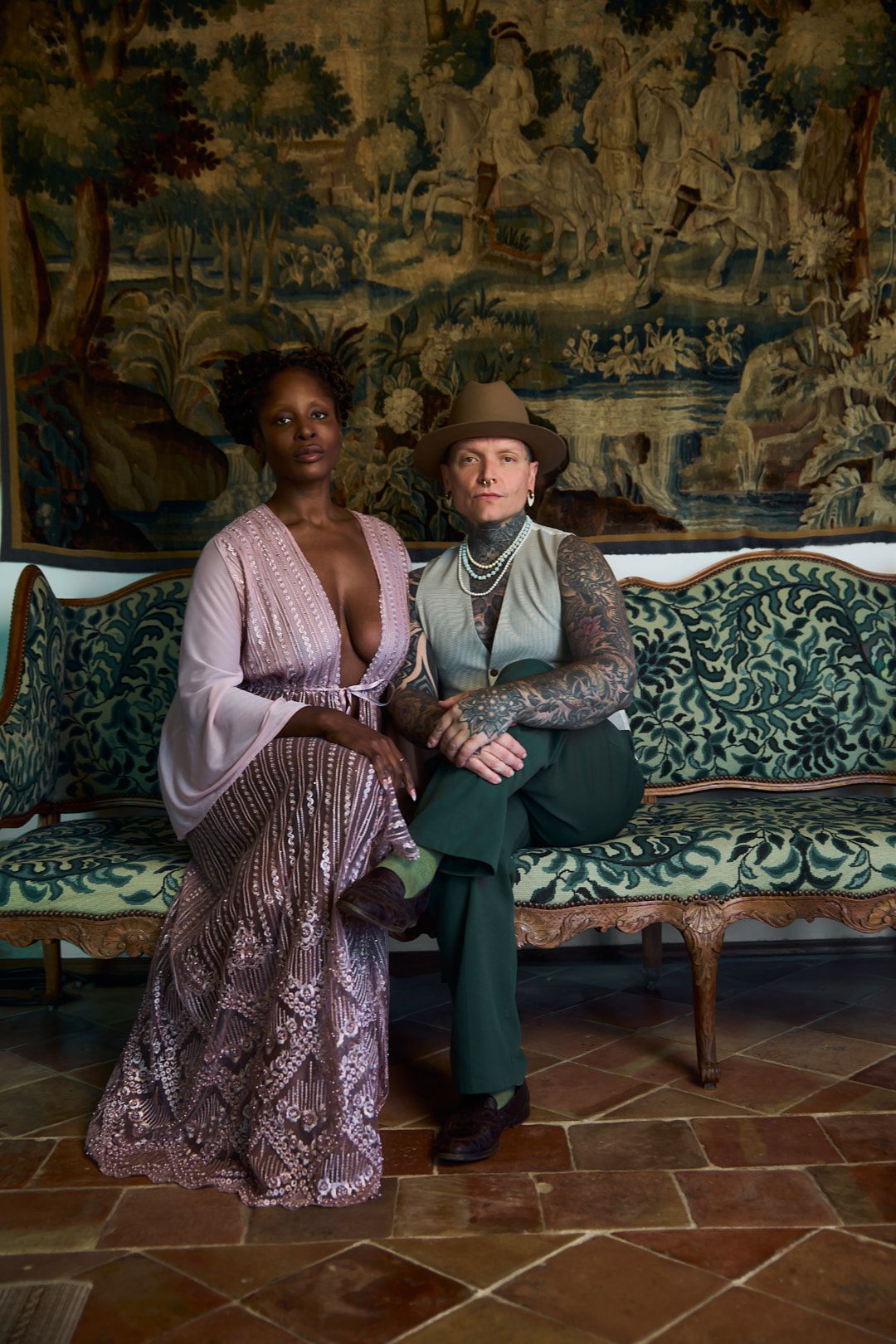 Two people sitting on a tiled floor against an ornate tapestry background in an elegant interior setting.