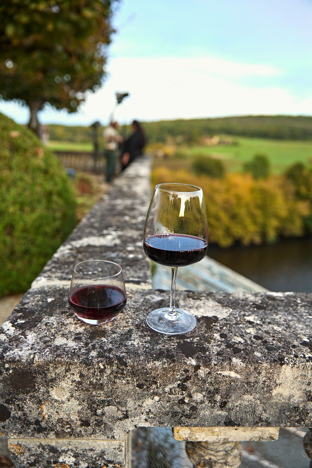 Glass of red wine sits on ancient stone wall overlooking scenic countryside view.