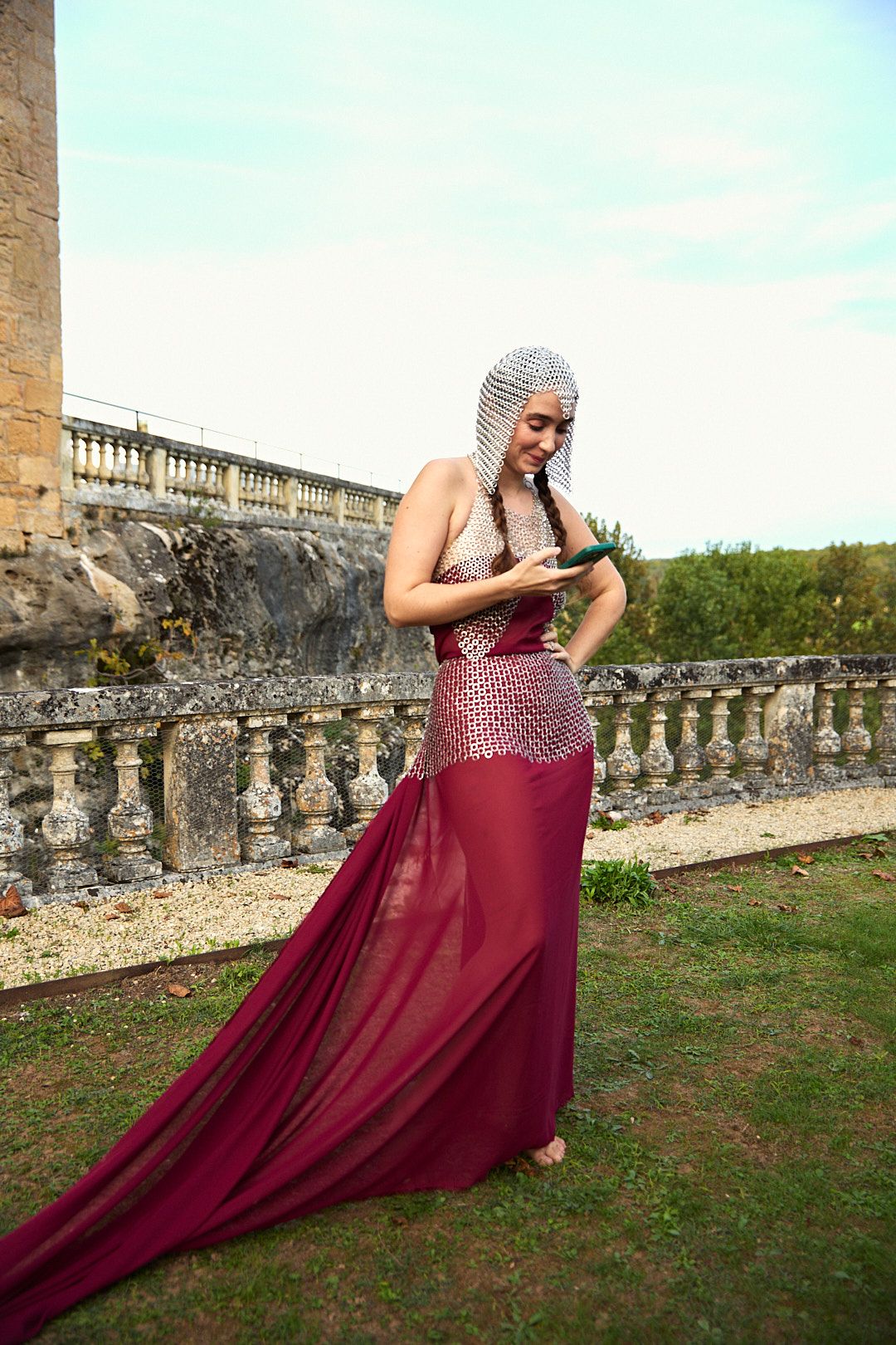 Person in flowing burgundy dress poses against stone balustrade in outdoor setting.