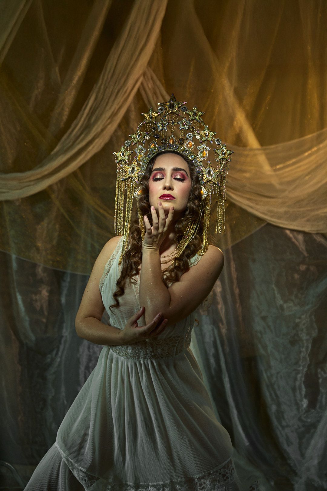 Artistic portrait with gold crown and flowing white dress against draped fabric backdrop.