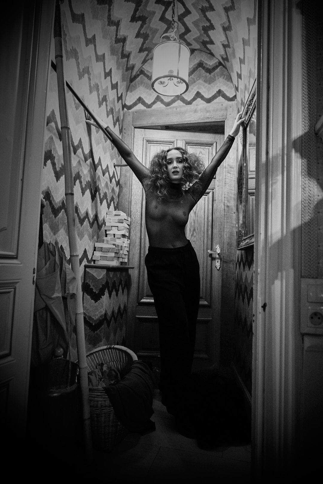 Artistic black and white photo of a figure posing with raised arms in a hallway with patterned wallpaper.