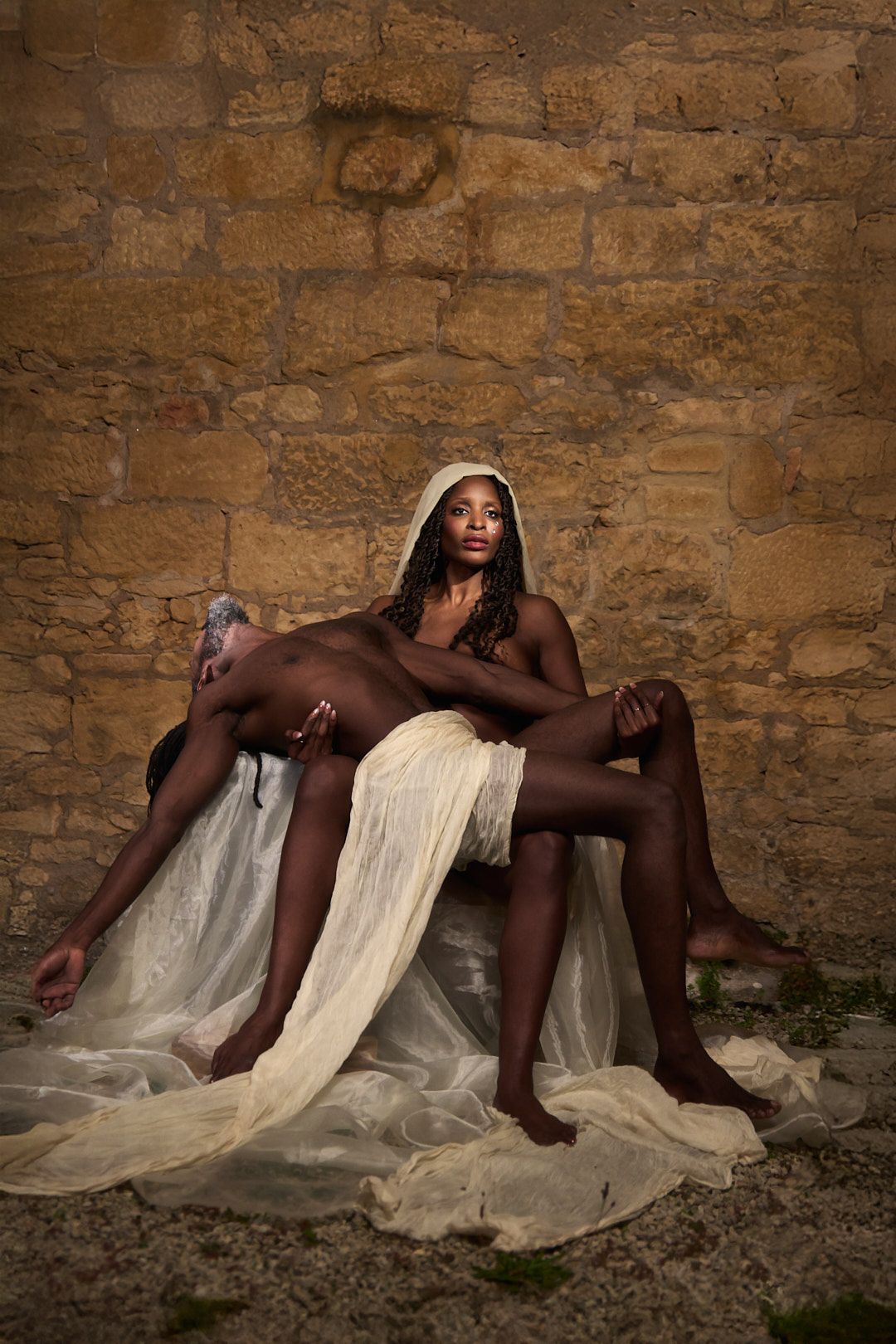 Artistic nude photo with white fabric draping against stone wall background.