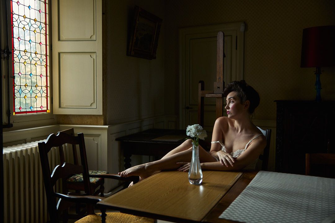 Moody interior scene with natural window light falling on figure at vintage table.