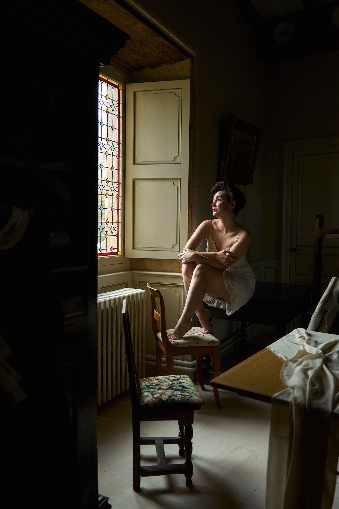 Artistic boudoir photoshoot in moody lighting showing a figure seated by a window in vintage interior.