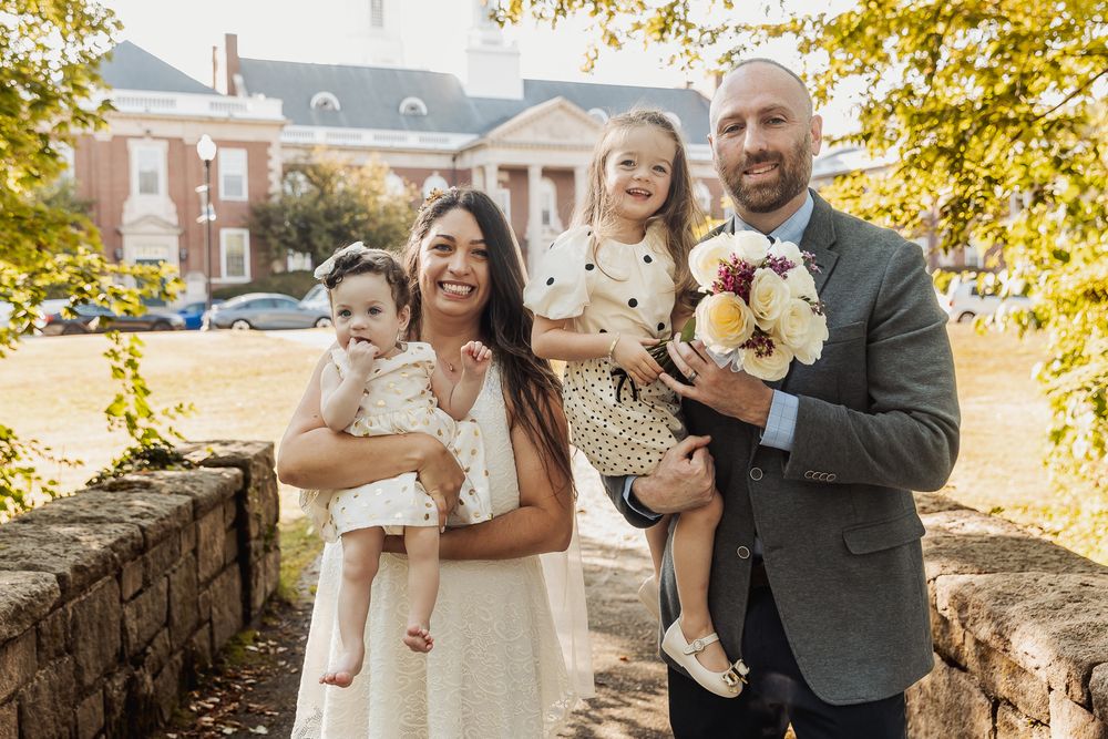 A happy couple poses outdoors on an autumn day with fall foliage and brick buildings in the background.