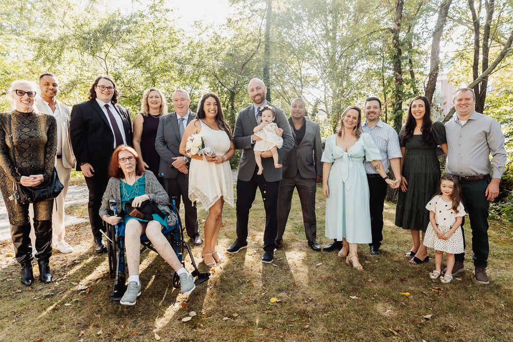 Large family group poses together outdoors in casual and formal attire during sunny woodland gathering.