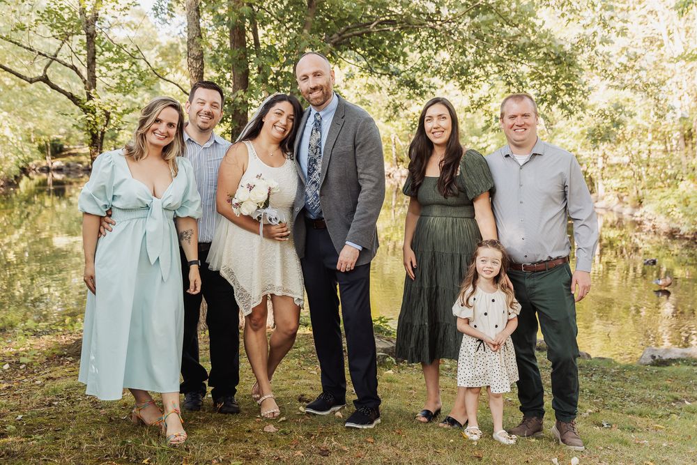 A group of people gather for an outdoor photo in a scenic park setting with trees and water in the background.