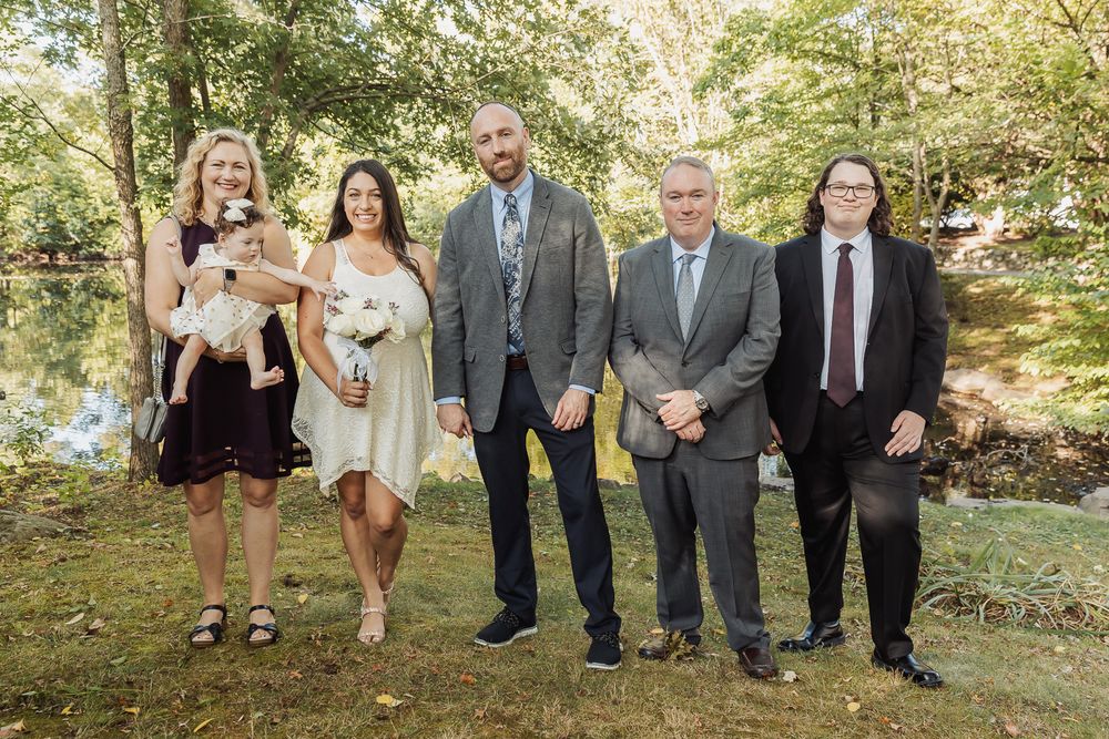 A group stands together outdoors in formal attire for a special occasion on a sunny day in a wooded setting.