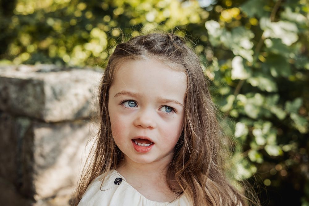 A young child with brown hair and blue eyes looks away from the camera while sitting outdoors in natural lighting.