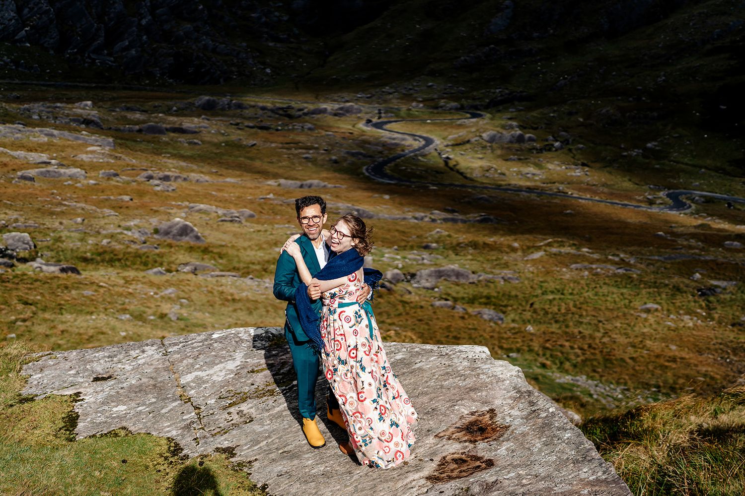 A couple in formal attire embrace on a rocky mountain outcrop surrounded by rugged wilderness landscape.