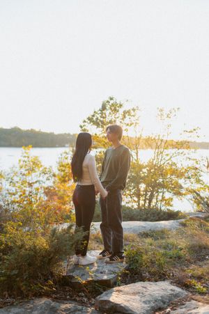 A couple stands together on a rocky outcrop overlooking a lake at sunset.