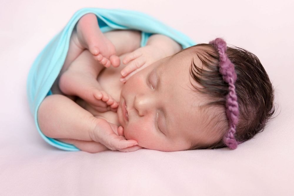 A sleeping newborn baby wearing a turquoise headband lies peacefully on a pink blanket.