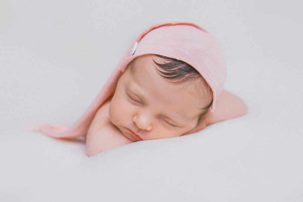 A sleeping newborn baby wearing a pink hat rests peacefully against a white background.