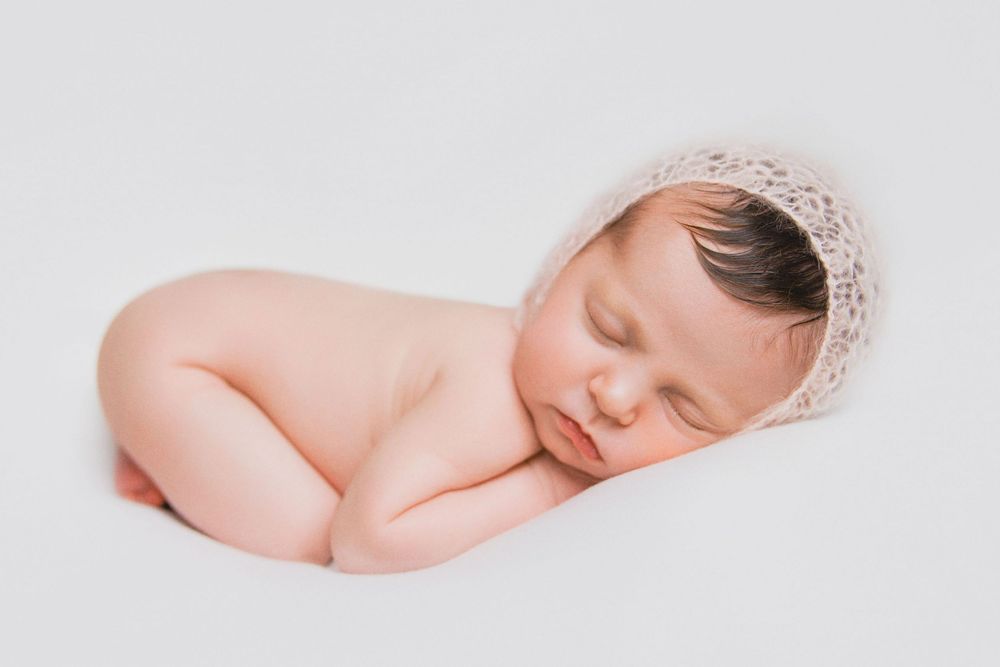 Peaceful sleeping newborn wearing delicate knit bonnet on white backdrop during professional photo session.