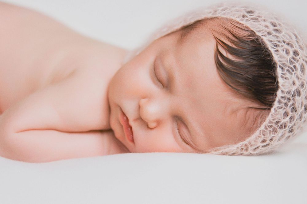 Sleeping newborn baby wearing delicate lace bonnet on soft pink background during professional photo session.